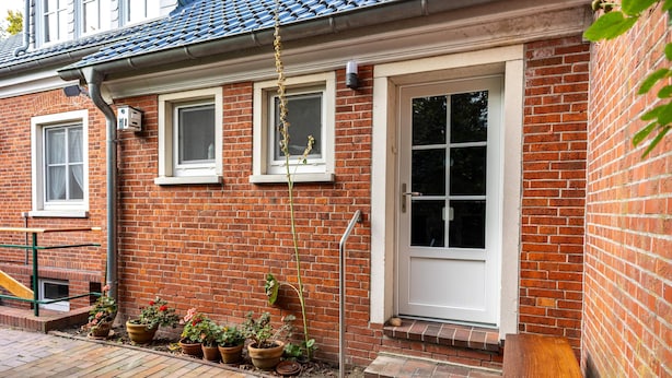 Ett hus byggt av tegel med en vit dörr, två fönster och en liten veranda med krukväxter.
