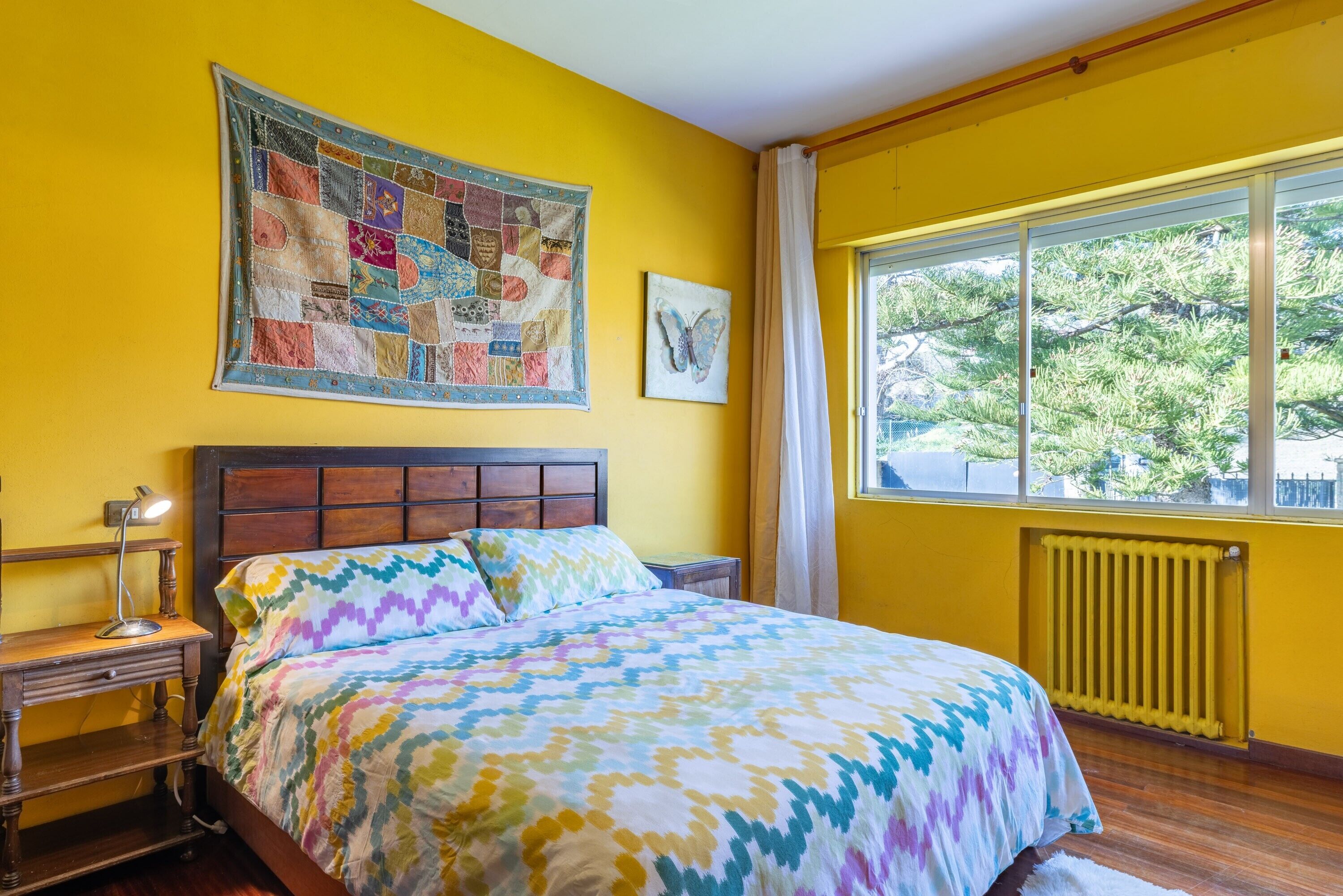 A bedroom with a wooden headboard, a quilt, a bedside lamp, a window with curtains, and a radiator.