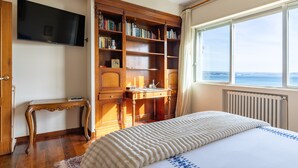 A bedroom with a bed, a wooden desk, a bookshelf, a television, and a window with a view.