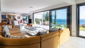 A modern living room with a large sofa, a coffee table, and a television. There is a sliding glass door leading to an outdoor area with a view of the sea.