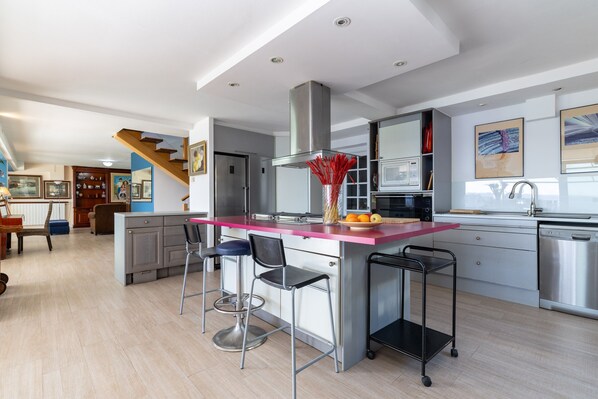 Une cuisine moderne dotée d’un îlot central, de tabourets de bar, d’appareils électroménagers en acier inoxydable et d’un escalier menant à un autre niveau.