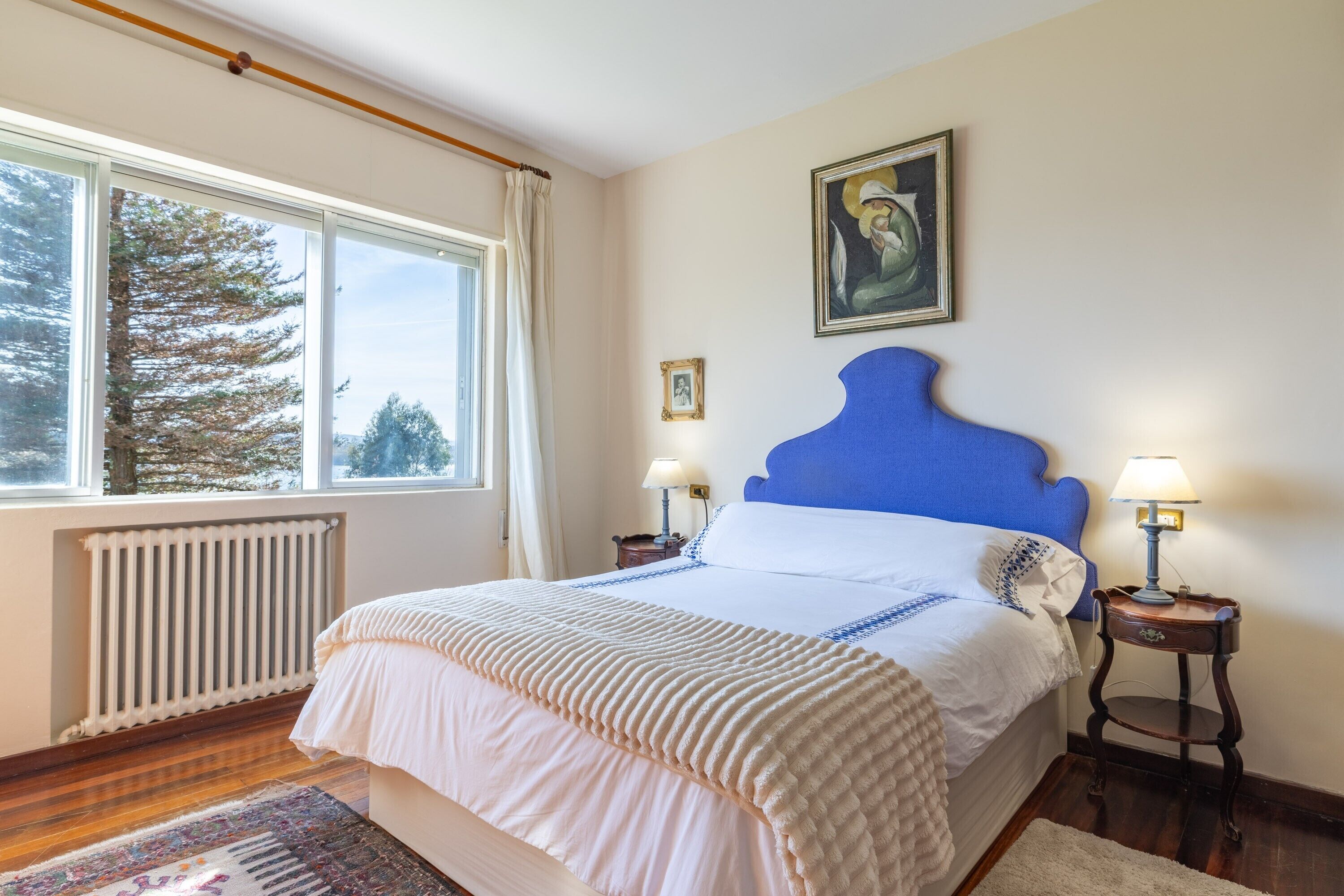 A bedroom with a large window, a radiator, a bed with a blue headboard, two bedside tables with lamps, and a framed picture on the wall.