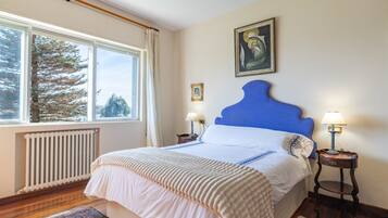 A bedroom with a large window, a radiator, a bed with a blue headboard, two bedside tables with lamps, and a framed picture on the wall.