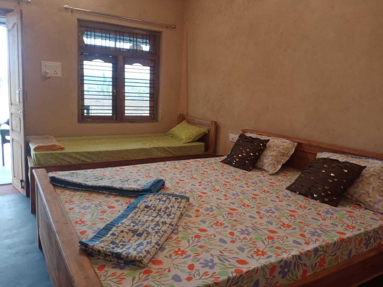 Una habitación con dos camas individuales, una con cubrecama verde y la otra con estampado floral. Hay una ventana con rejas, y las paredes están pintadas de un color claro.
