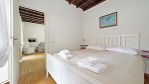 Een slaapkamer met een bed, nachtkastje, bijzettafeltje, schilderij en een deur naar een andere kamer.