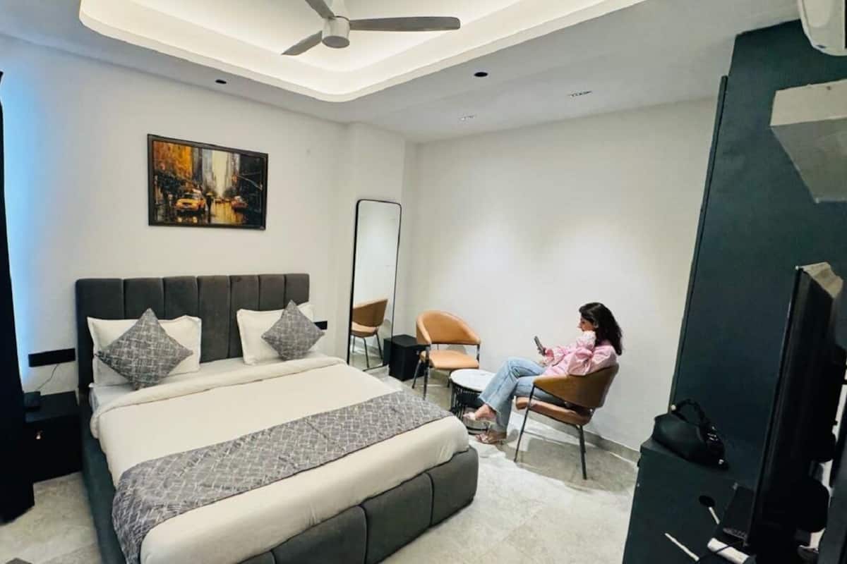 A modern bedroom with a large bed, a ceiling fan, a painting on the wall, and a person sitting on a chair using a mobile device.
