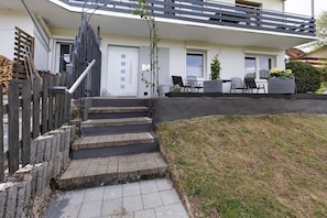 A residential building with a front entrance, steps leading to the door, a small outdoor seating area with chairs and a table, and a garden area with a hedge and a potted plant.