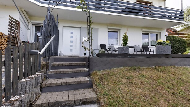 A residential building with a front entrance, steps leading to the door, a small outdoor seating area with chairs and a table, and a garden area with a hedge and a potted plant.