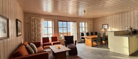 A spacious living room with a wooden ceiling, a dining area with a kitchen counter, and large windows.