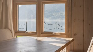 Habitación con un amplio ventanal que ofrece vistas a montañas y un lago. Hay una mesa de madera y un banco acolchado.