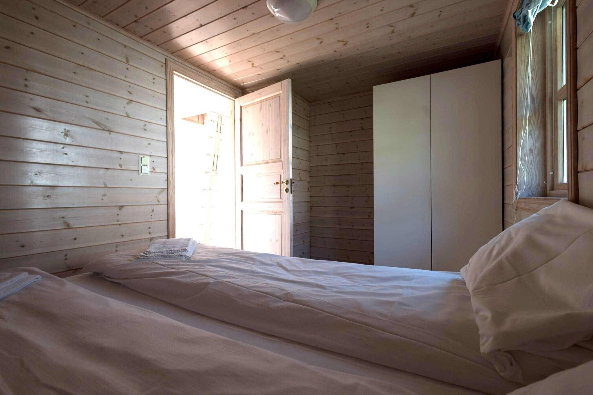 Una habitación pequeña con cama individual, paredes y techo de madera. Incluye un armario y una ventana con cortinas.