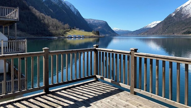Una terraza de madera con barandilla que da a un lago tranquilo, rodeado de montañas nevadas.