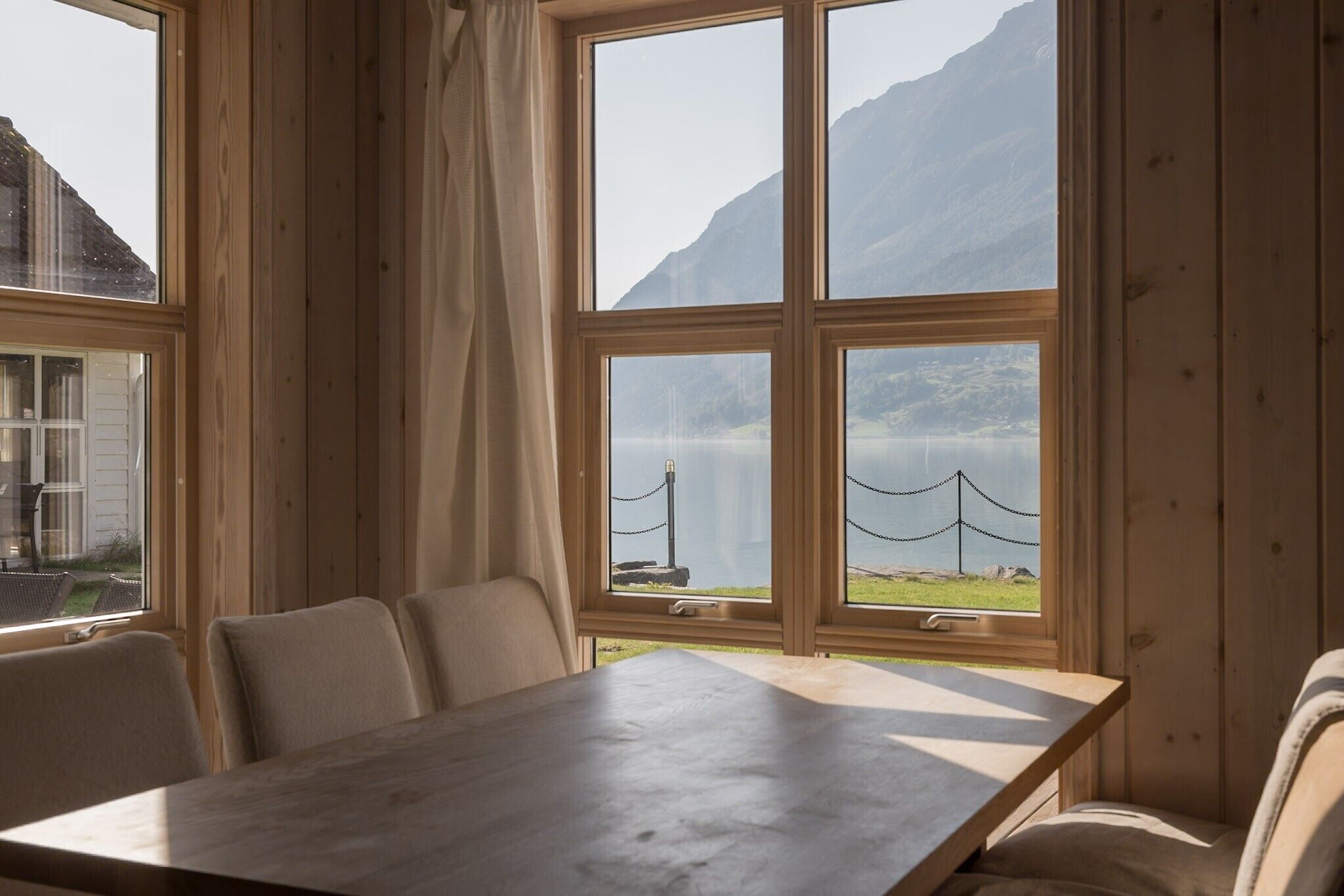 A wooden interior with a table and chairs, large windows with a view of mountains and a body of water.