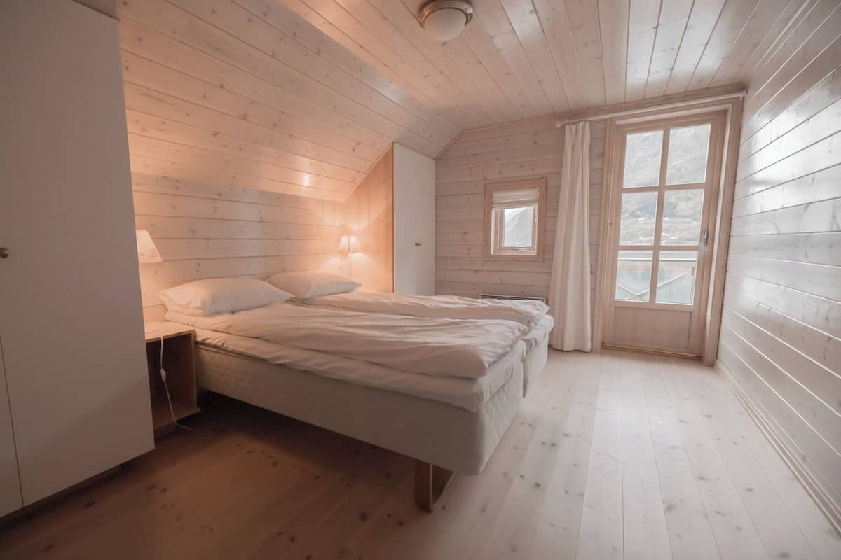 A bedroom with a wooden slat ceiling, a bed with white bedding, a nightstand, and a window with curtains.