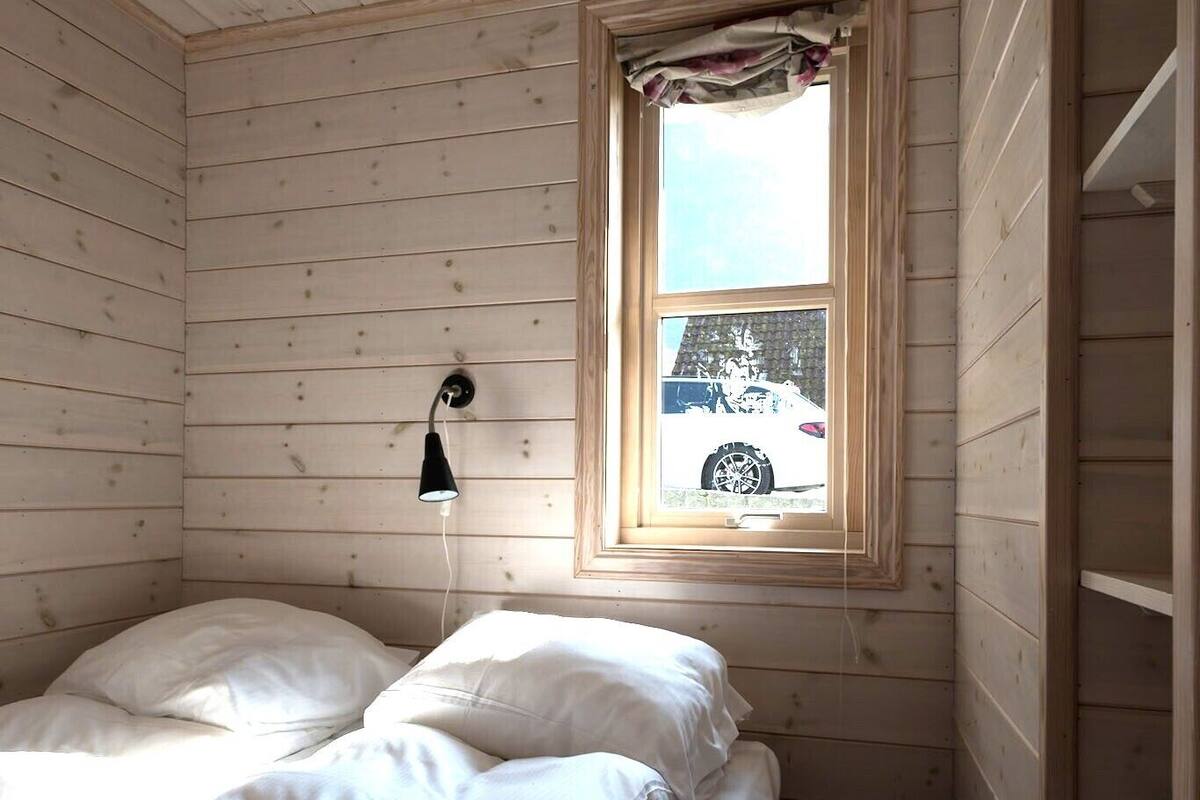 A small, wooden room with a single bed, a window with a view of a car outside, and a wall-mounted light fixture.