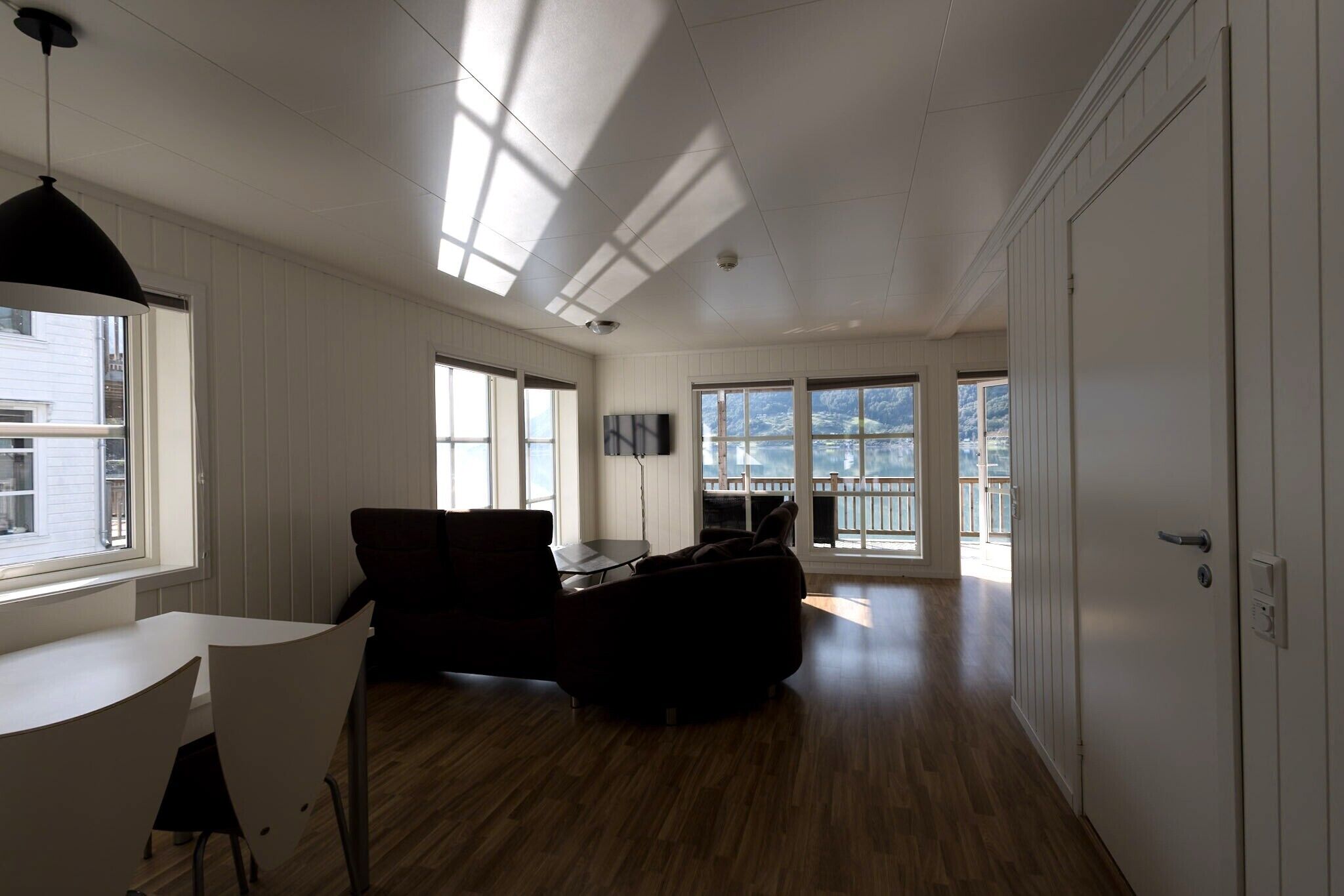 A spacious living area with a wooden floor, a dining table with chairs, a sofa, and a TV. Large windows allow natural light and views of the outdoors.