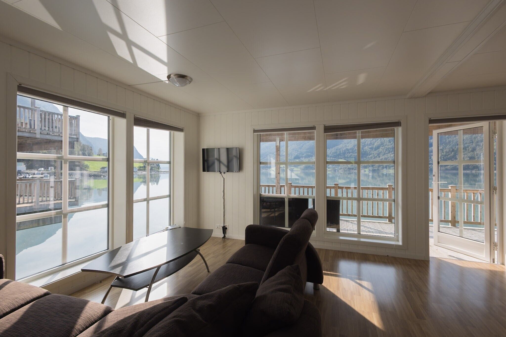 Ein geräumiges Zimmer mit großen Fenstern und Blick auf ein Ufer sowie Berge. Es gibt ein Sofa, einen Tisch und einen an der Wand befestigten Flachbildfernseher.