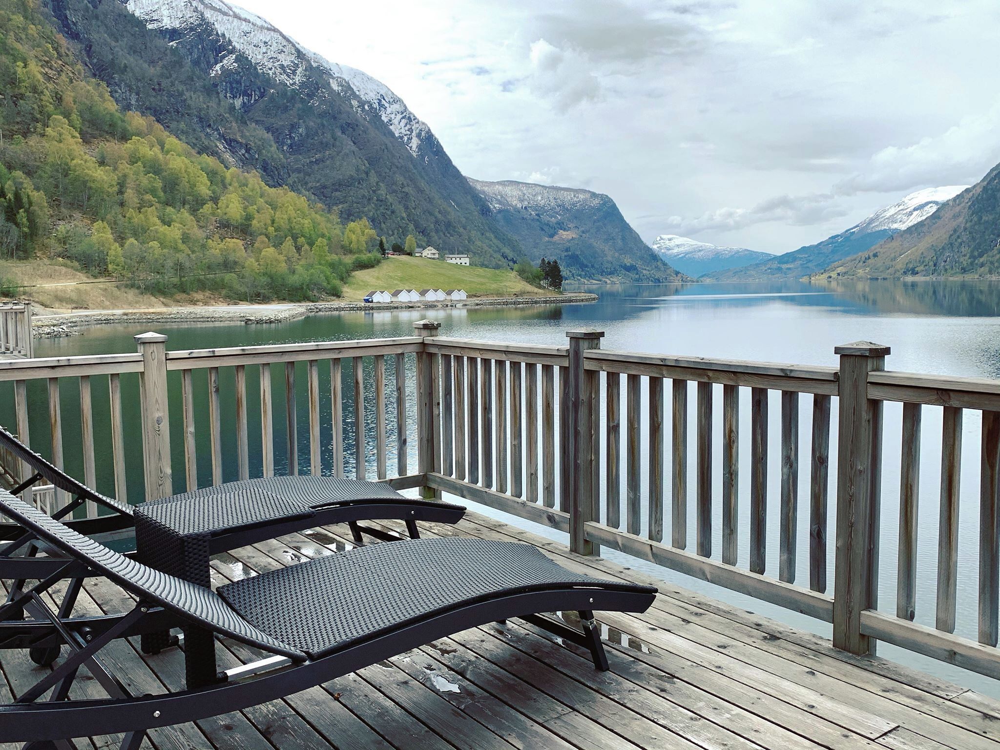 A wooden deck with two lounge chairs overlooking a lake, surrounded by mountains and a row of cabins.