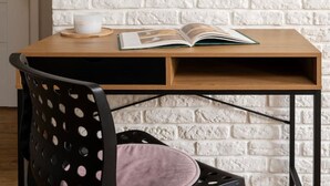 Een kamer met een witte bakstenen muur, een houten bureau met een boek en een kopje, en een zwarte stoel met een roze kussen.