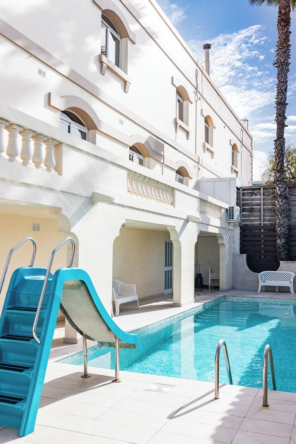 Un espace piscine avec un toboggan bleu, un bâtiment blanc orné d’arches et un palmier.