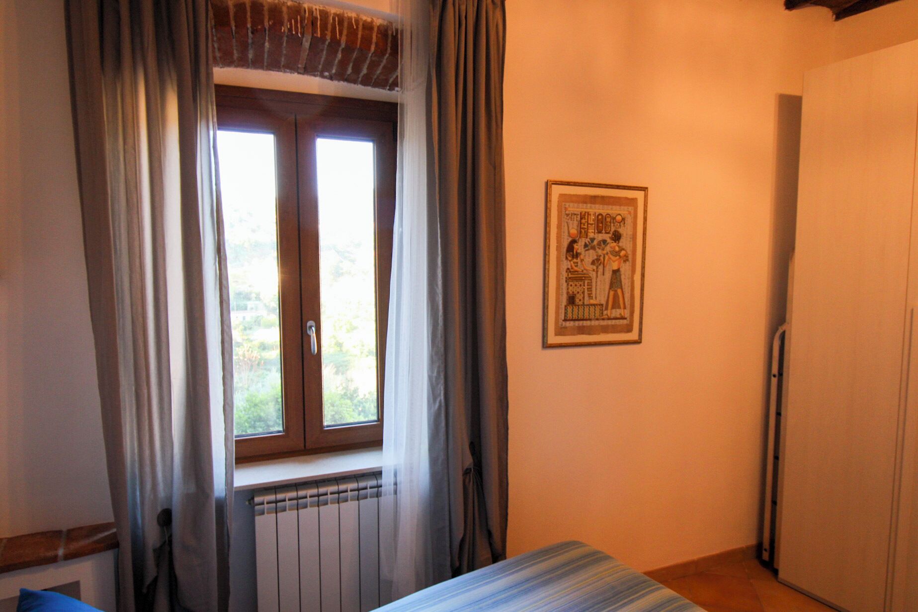 Ein Schlafzimmer mit Fenster, Heizkörper, Bett und Tür, sowie einem gerahmten Bild an der Wand.