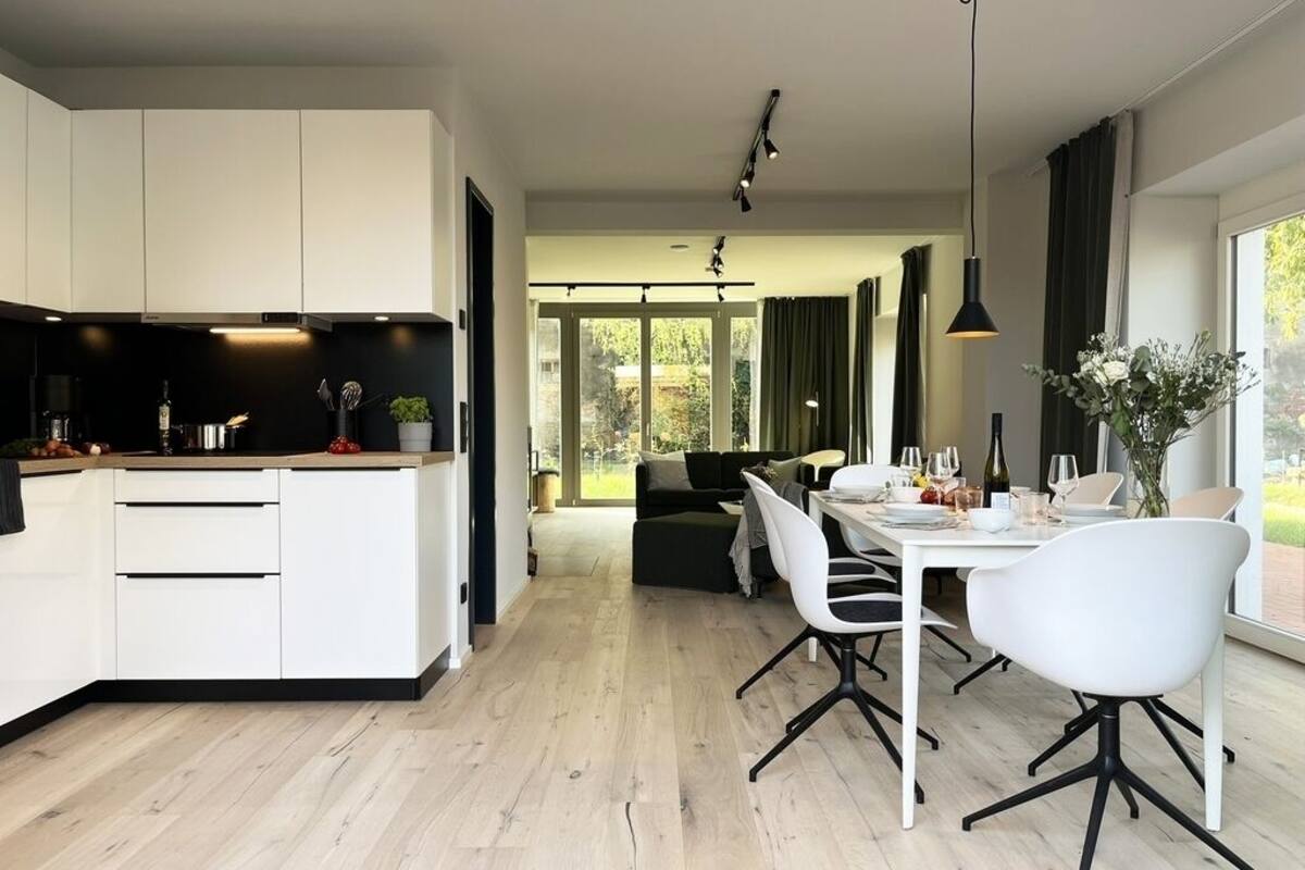 A modern kitchen with white cabinets, a dining area with a white table and chairs, and a view of the outdoors through large windows.