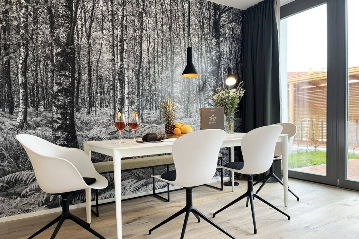 A dining area with a white table and four chairs, a forest-themed wallpaper, a black pendant light, and a window with a view of a building.