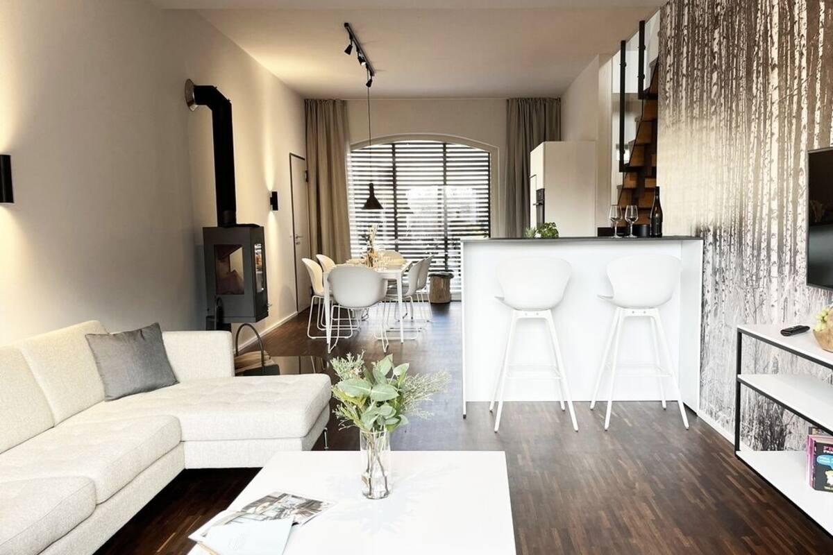 A modern living room with a white sofa, a coffee table with a vase, a dining area with a table and chairs, and a kitchen area with a bar and stools.