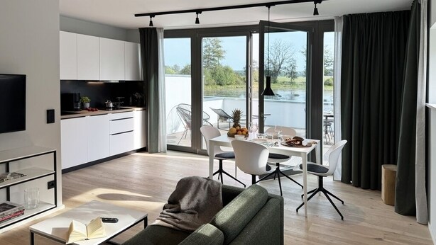 A modern living room with a sofa, a coffee table, and a TV. There is a dining area with a table and chairs, and a kitchen with cabinets and appliances. The room has large windows with curtains. - Resort Schloss Reinharz (Bad Schmiedeberg)