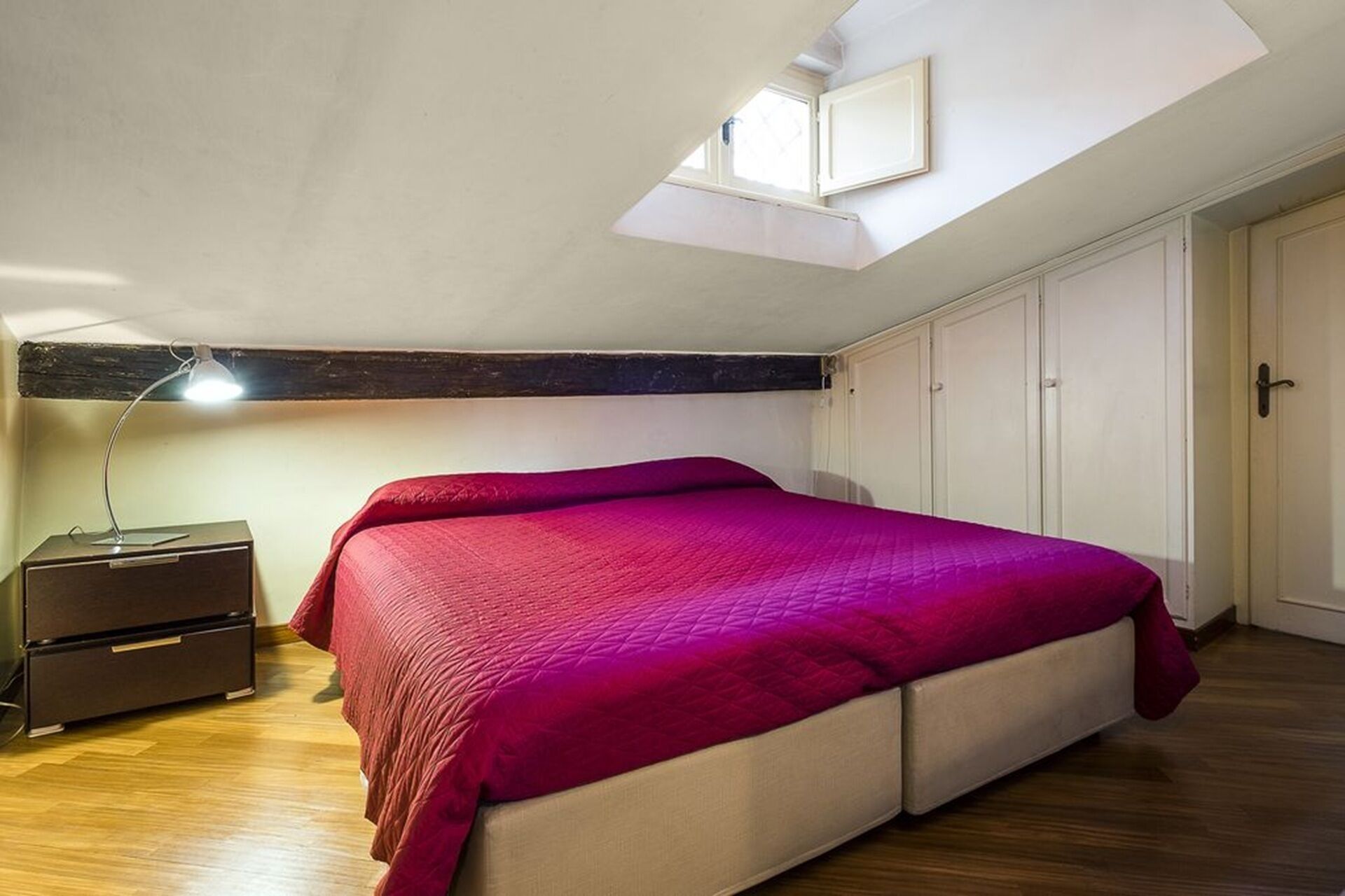 A bedroom with a skylight, a bed with a red blanket, a nightstand, and a wardrobe.