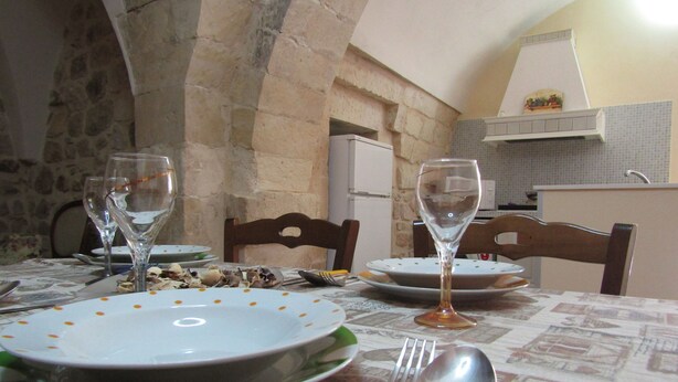 A dining table set with plates, cutlery, and wine glasses, with stone walls and a kitchen in the background. - The Courtyard Room Fishing (Scicli)