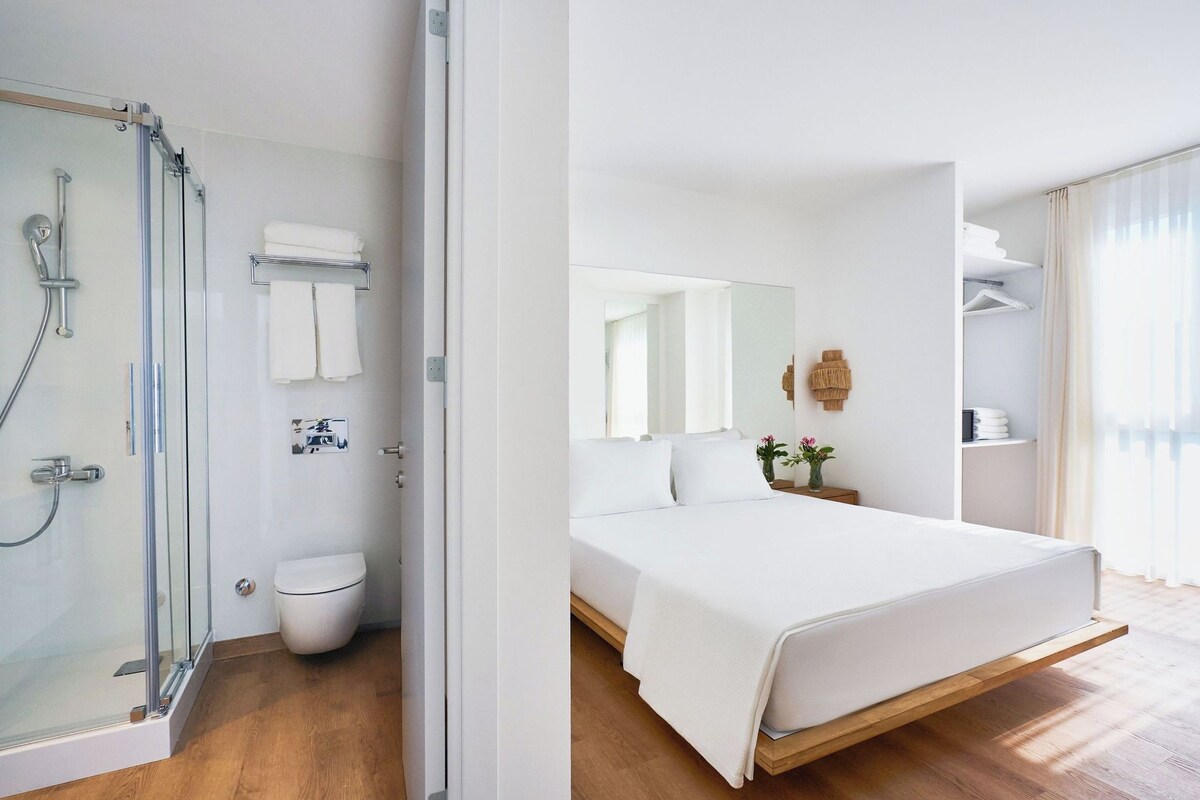 Une chambre d’hôtel moderne dotée d’une douche à parois de verre, de toilettes blanches, d’un lit avec un cadre en bois et d’un grand miroir.