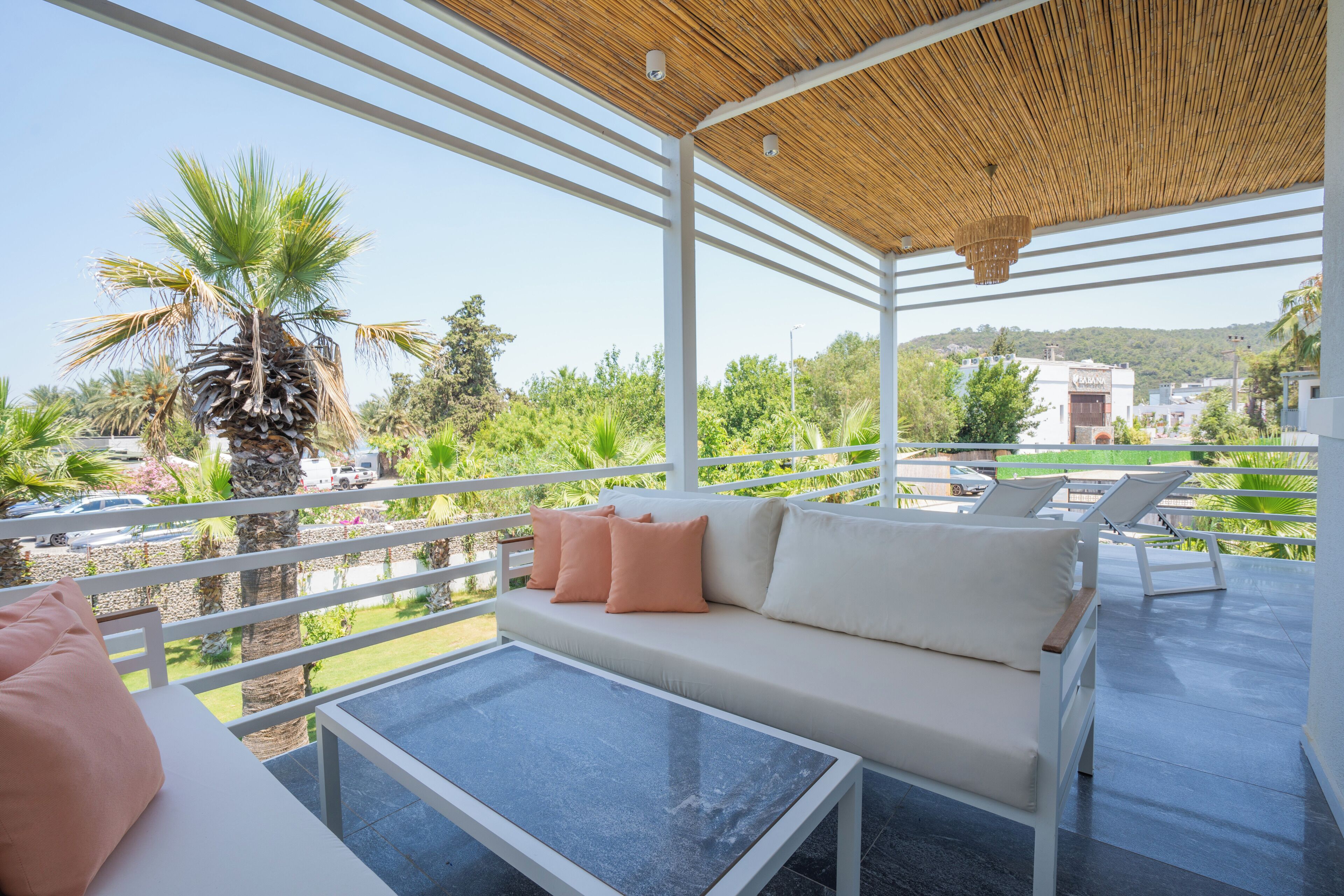 Un espace extérieur aménagé, abrité et doté d’un canapé et de fauteuils, offrant une vue sur un paysage luxuriant, avec des palmiers et un bâtiment situé au loin.