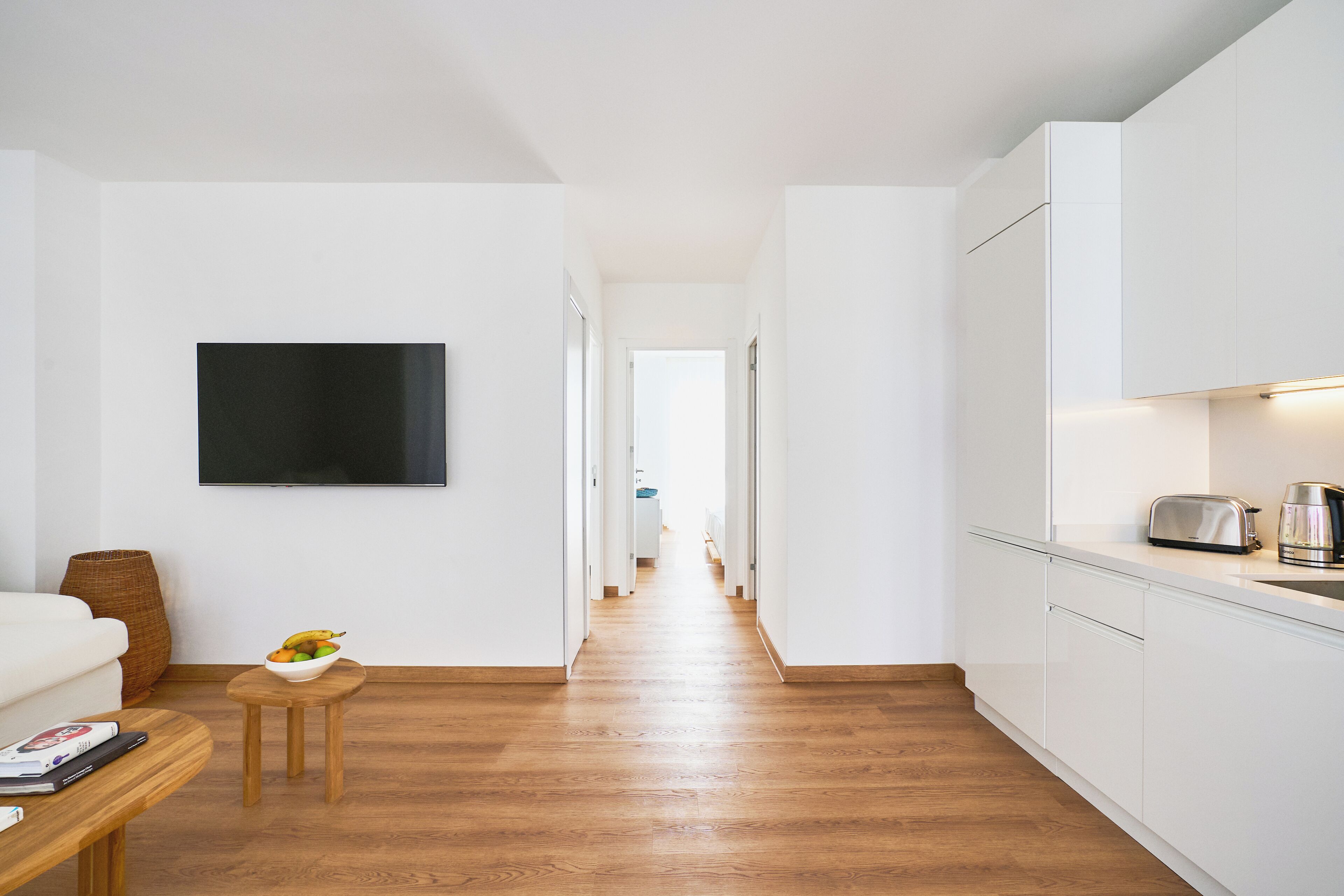 A modern kitchen with white cabinets, a wooden floor, and a small table with a bowl of fruit.