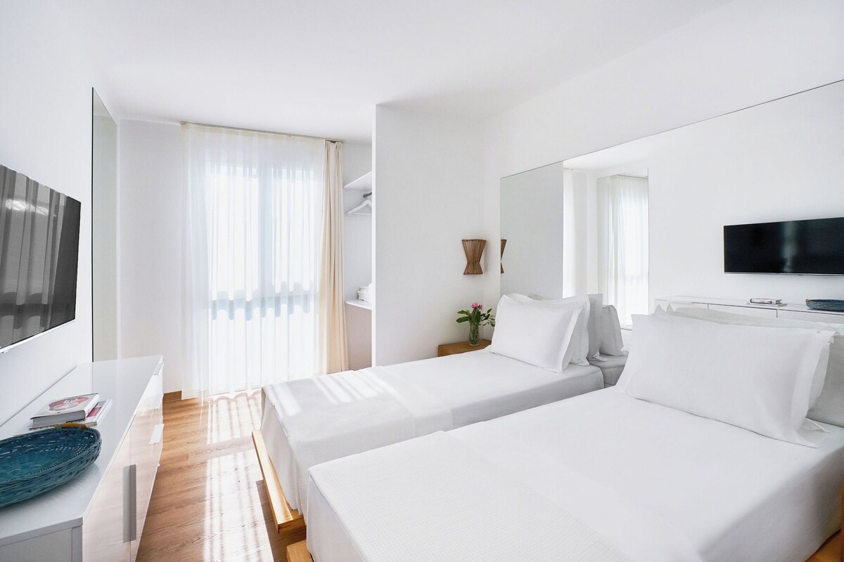 A hotel room with two beds, a TV, a desk, and a window with curtains.