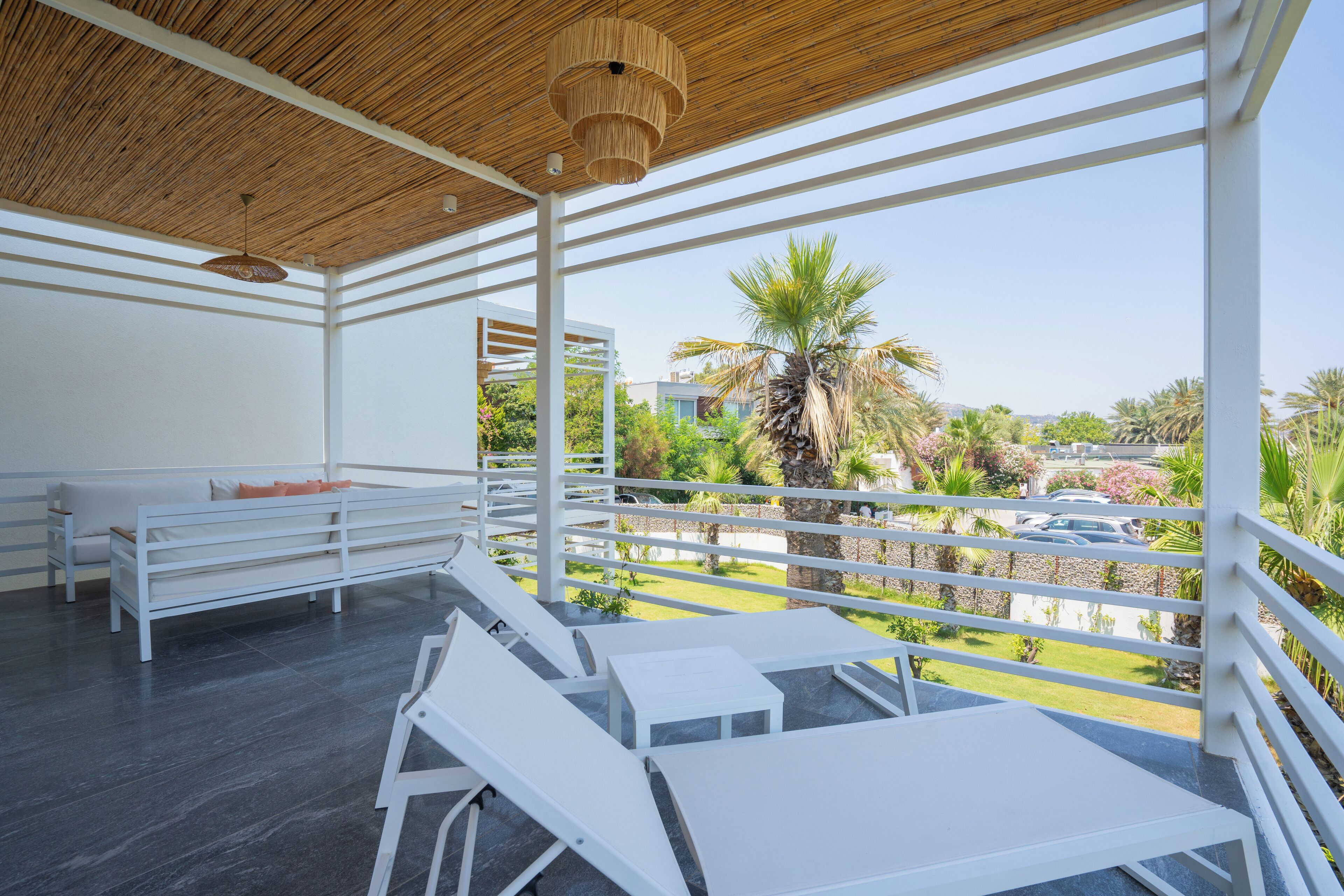 A modern outdoor lounge area with white furniture, a wooden ceiling, and a view of a landscaped garden and palm trees.
