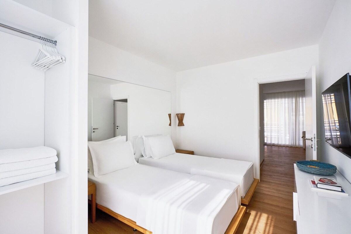 Une chambre d’hôtel avec deux lits, une télévision, un miroir, un plancher en bois et une porte donnant sur une autre chambre.