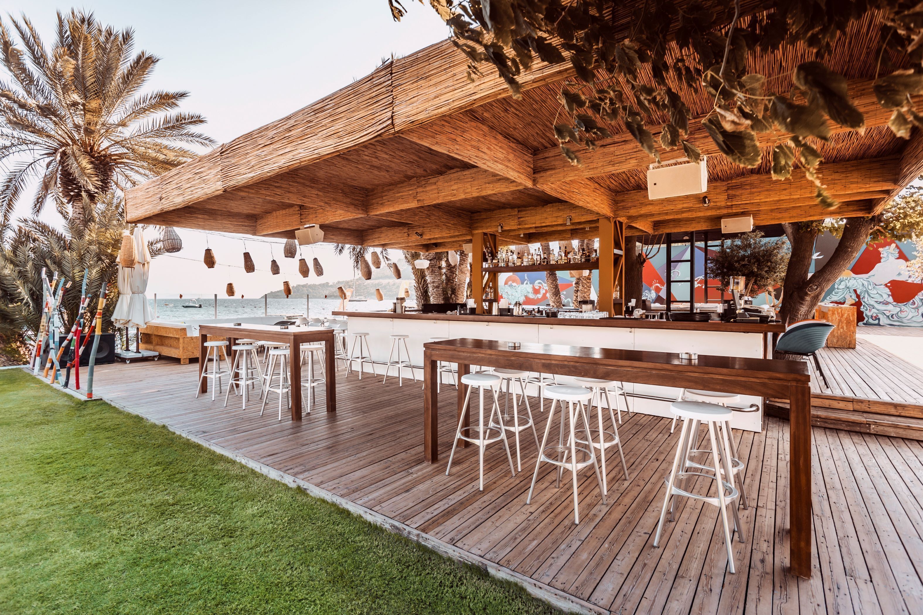 Una terraza de madera con zona de bar, taburetes blancos y vista al mar.