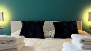 Een net opgemaakt bed met witte lakens en twee zwarte kussens, een blauwgroene wand en twee wandlampen.