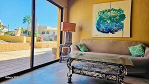 Sala de estar con sofá, mesa de centro y lámpara. Amplia ventana con vista a palmeras y edificios.