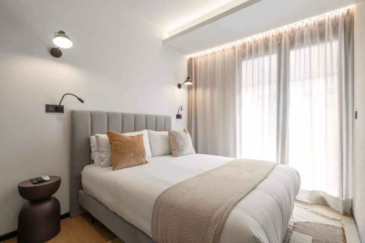 A modern bedroom with a large bed, two bedside lamps, and a view of the outdoors through sheer curtains.