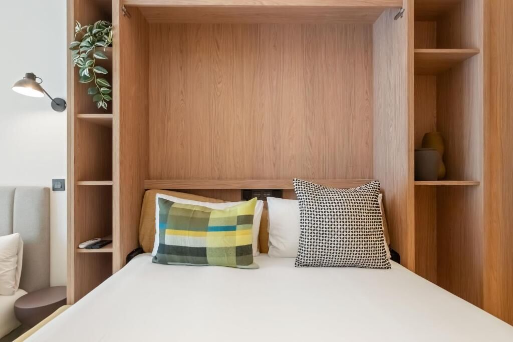 Un dormitorio pequeño con cabecera de madera, dos almohadas, una lámpara de noche y una pequeña estantería con una planta.