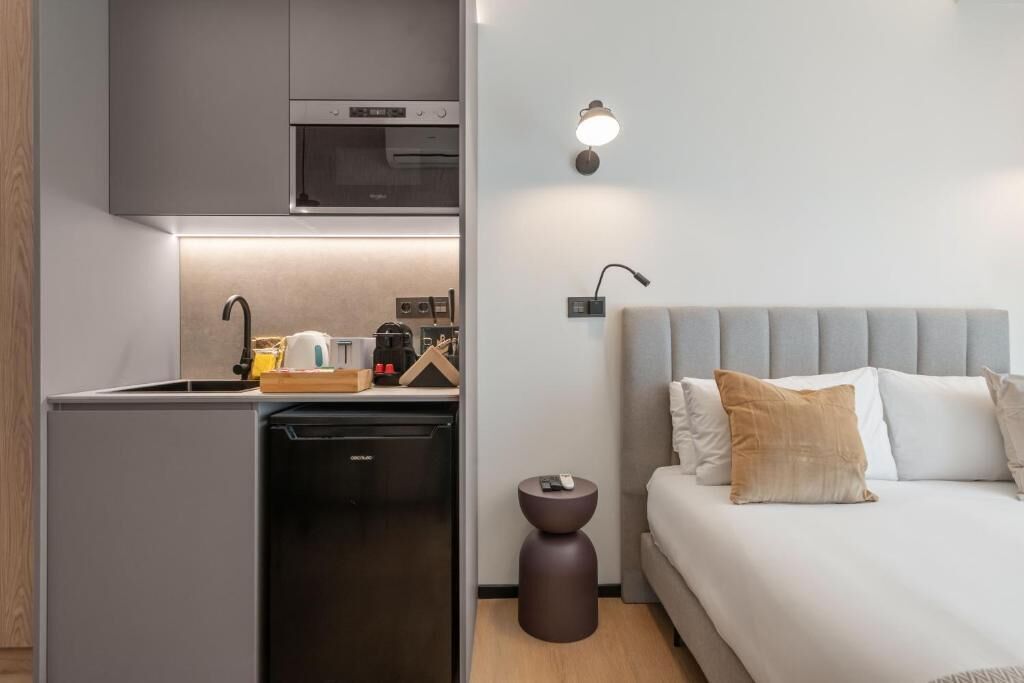 Habitación de hotel pequeña con kitchenette, una cama y un cabecero gris, y una mesita de noche con lámpara.