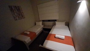 Ein kleiner, schummrig beleuchteter Raum mit zwei Einzelbetten, jeweils mit einem Kopfkissen und einem zusammengerollten Handtuch.