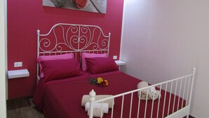 A bedroom with a red wall, a white metal bed frame, two pink pillows, and a single red rose on the bed. - The Red Room Courtyard (Scicli)