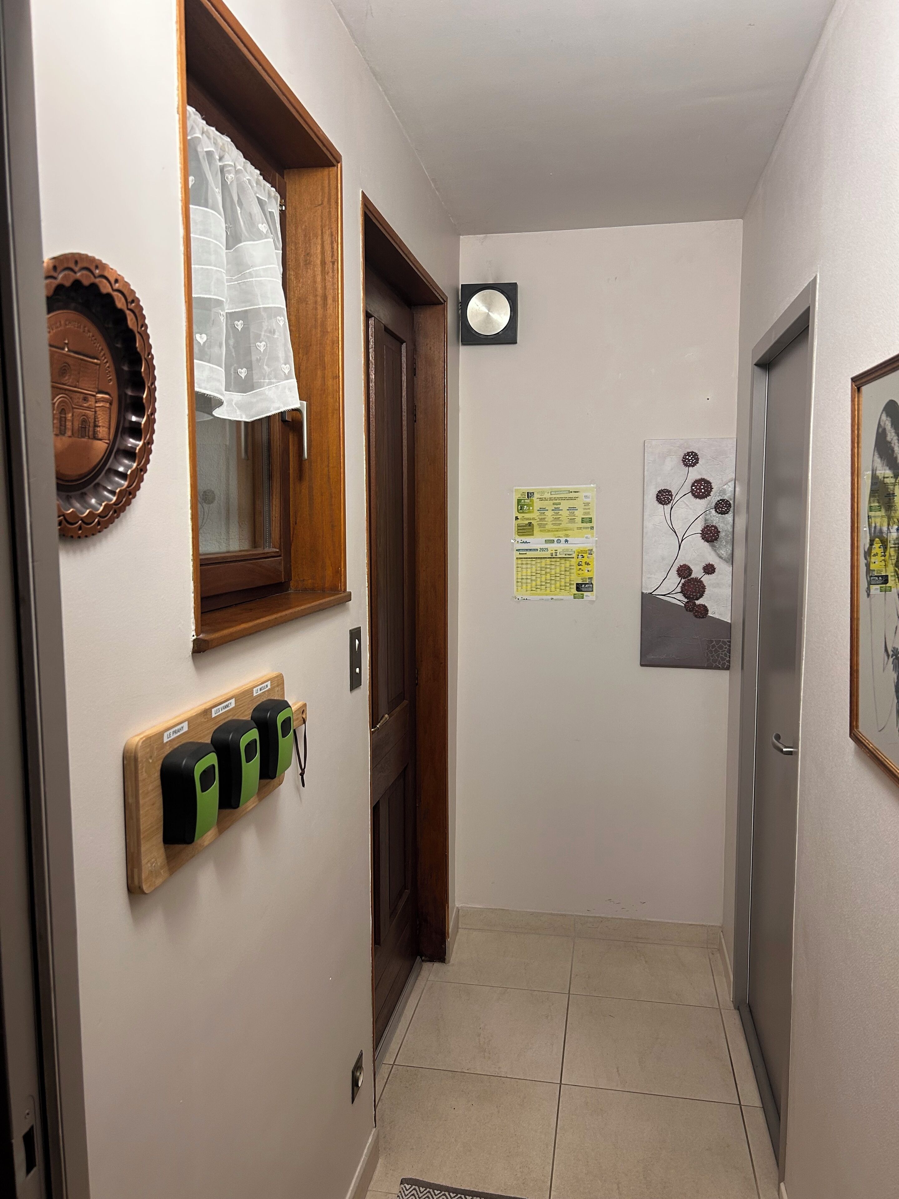 Een smalle gang met een houten deur, een raam met gordijn, een lichtschakelaar op de muur en een klein prikbord met papieren.