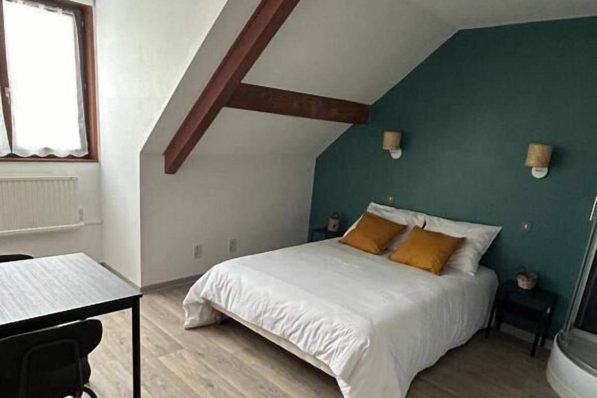 A bedroom with a sloped ceiling, a bed with white bedding and two orange pillows, a small bedside table, a nightstand, a window with a curtain, and a radiator.