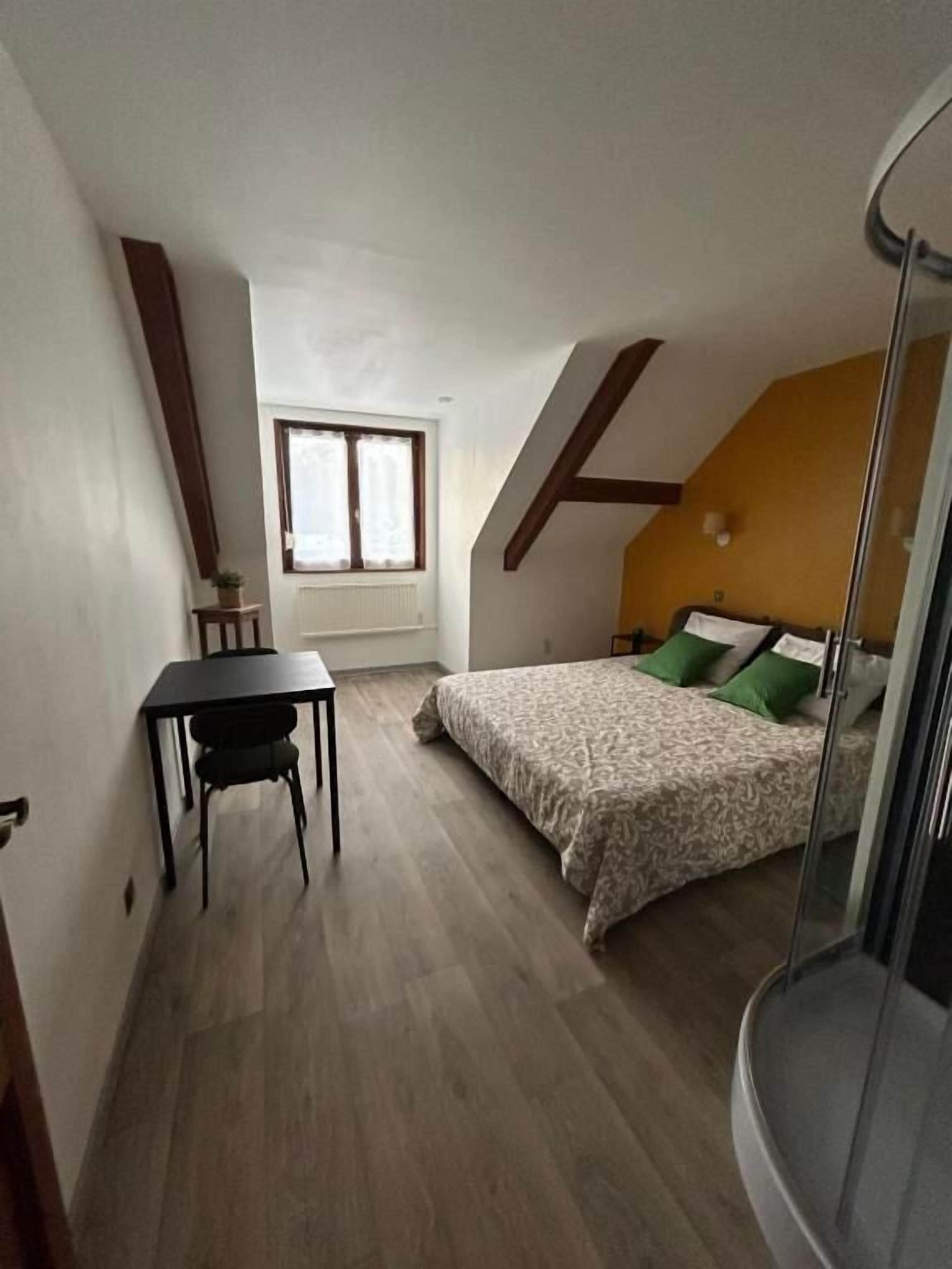 Een klein appartement met één kamer en een schuine kap, een bed met groene kussens, een tafeltje met een stoel en een raam met gordijnen.
