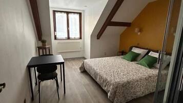 Een klein appartement met één kamer en een schuine kap, een bed met groene kussens, een tafeltje met een stoel en een raam met gordijnen.