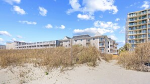 A beachfront hotel with multiple floors, a sandy path leading to the entrance, and a clear sky. - Dog Friendly-Windy Village-Oceanfront-Sleeps6-2br/2ba-Full Kitchen-Free Parking (North Myrtle Beach)